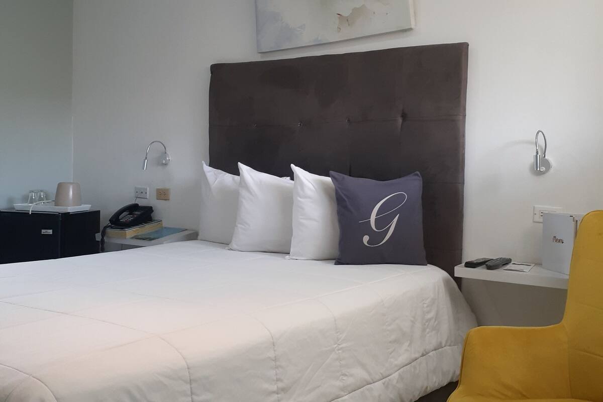 Hotellihuone, jossa on suuri sänky, yöpöytä lampun kanssa, tuoli ja pieni työpöytä, jossa on puhelin ja kansio.