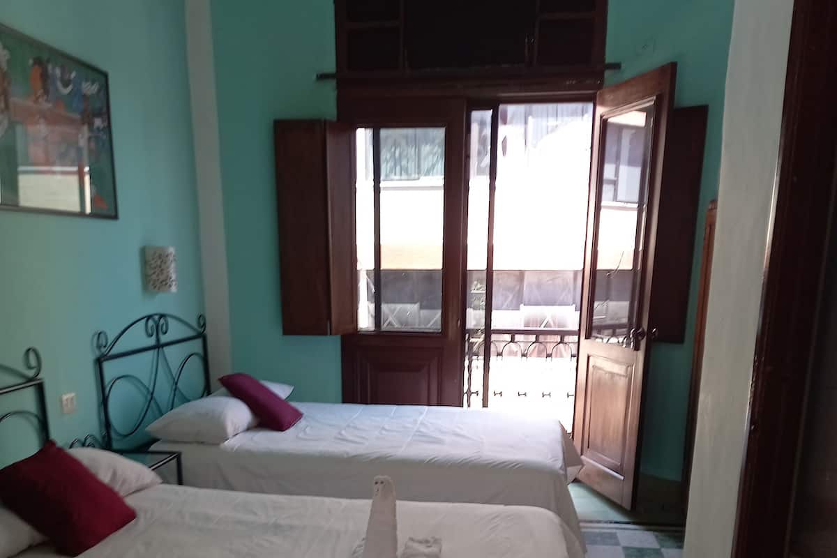 A hotel room with two beds, a checkered floor, a balcony with a view, and a door leading outside.