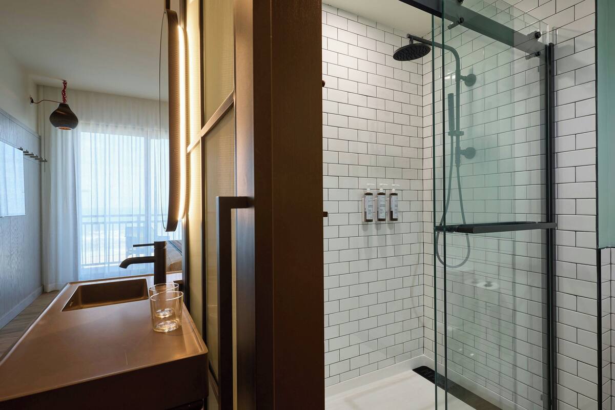 A modern bathroom with a glass-enclosed shower, a wooden vanity with a sink and glass, and a large window with sheer curtains.
