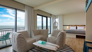Habitación de hotel con un ventanal grande que ofrece vistas al océano, una cama con cubrecama estampado, dos sillones color beige, una mesita pequeña con un objeto decorativo y una alfombra estampada.