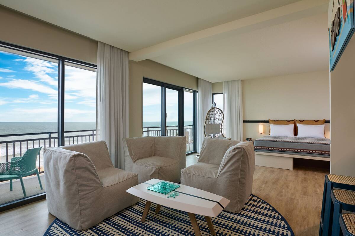 A hotel room with a large window offering a view of the ocean, a bed with a patterned bedspread, two beige armchairs, a small white table with a decorative item, and a patterned rug.