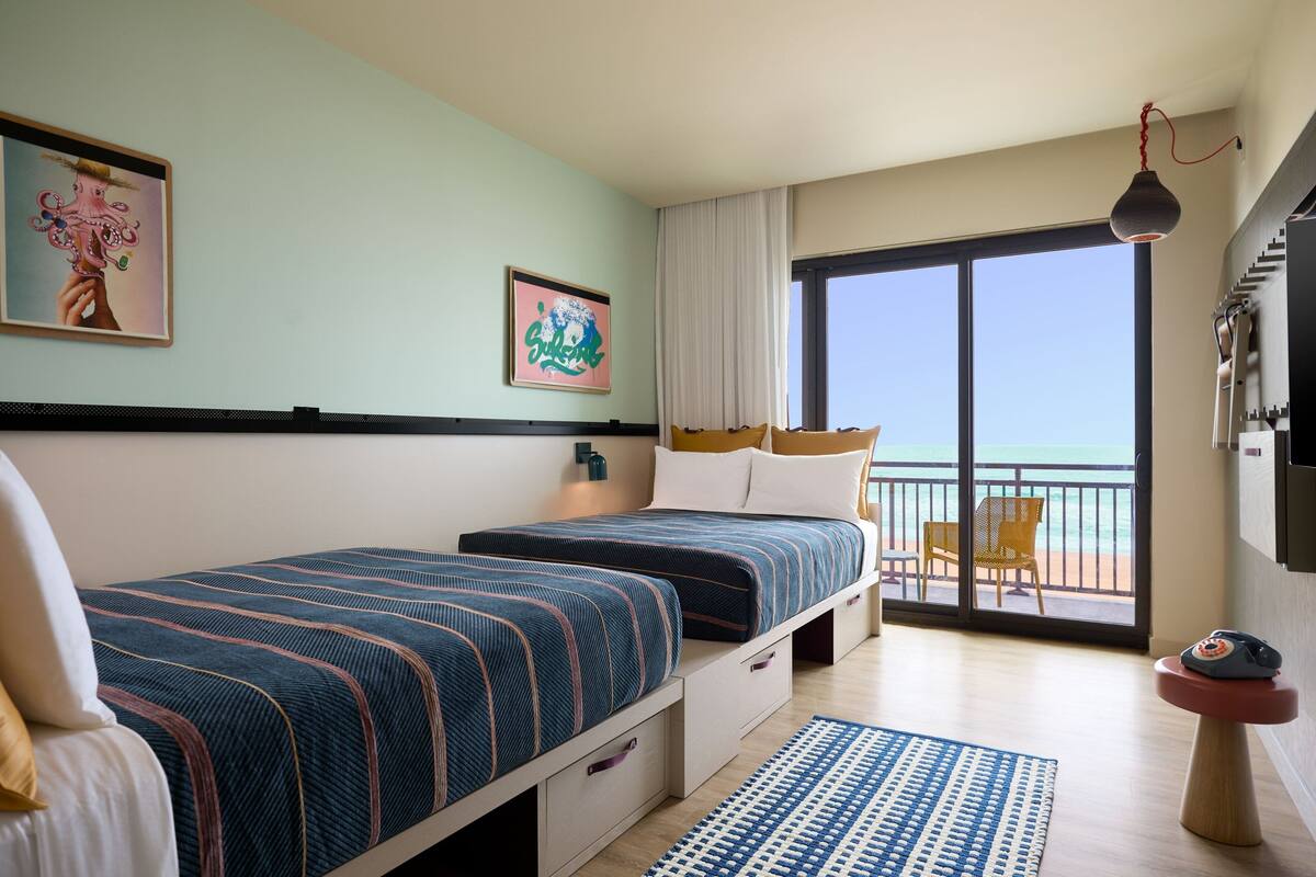 A hotel room with two beds, a balcony with a chair, and a view of the sea.