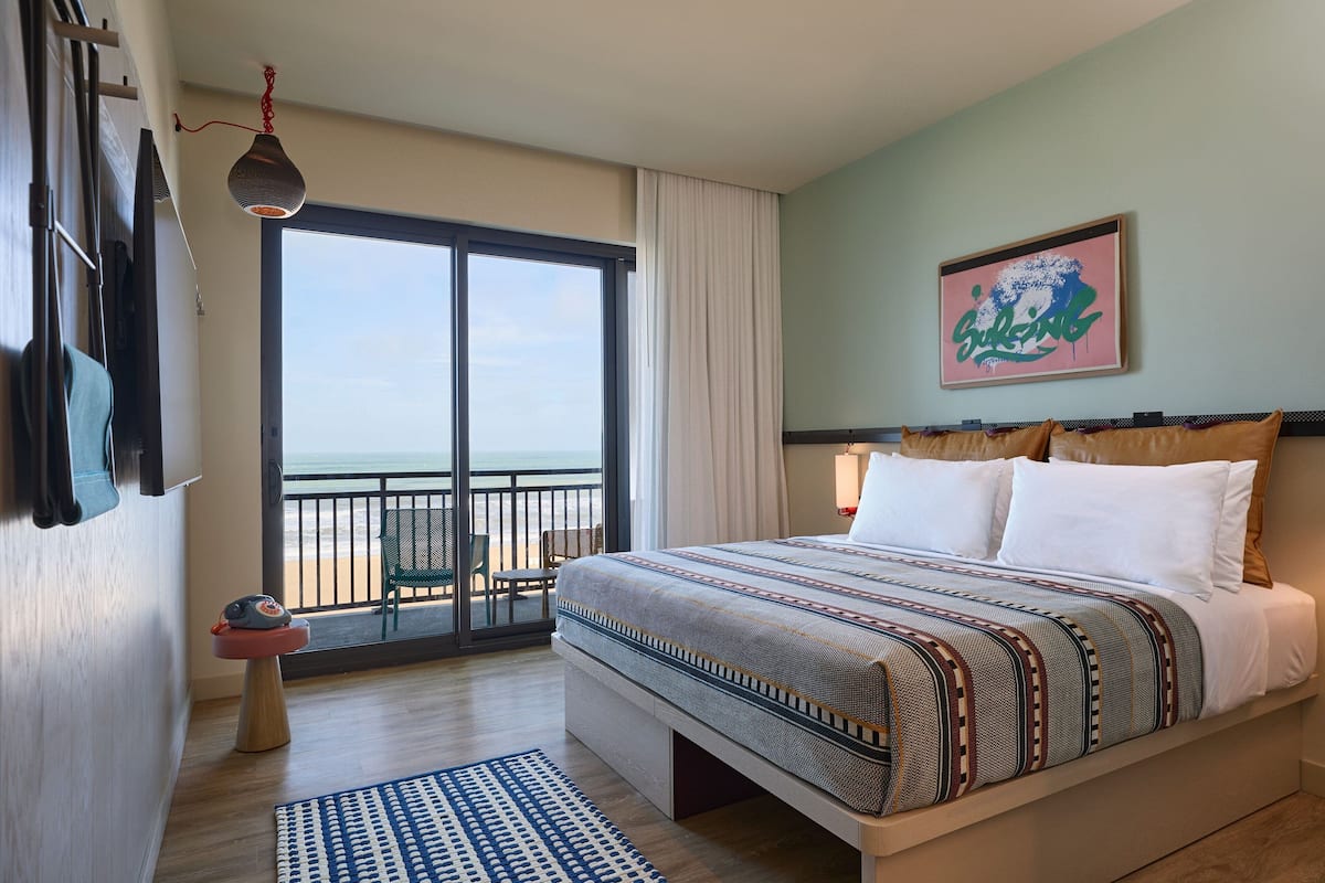 A hotel room with a large bed, a view of the beach, a balcony with a chair, and a small table with a toy.