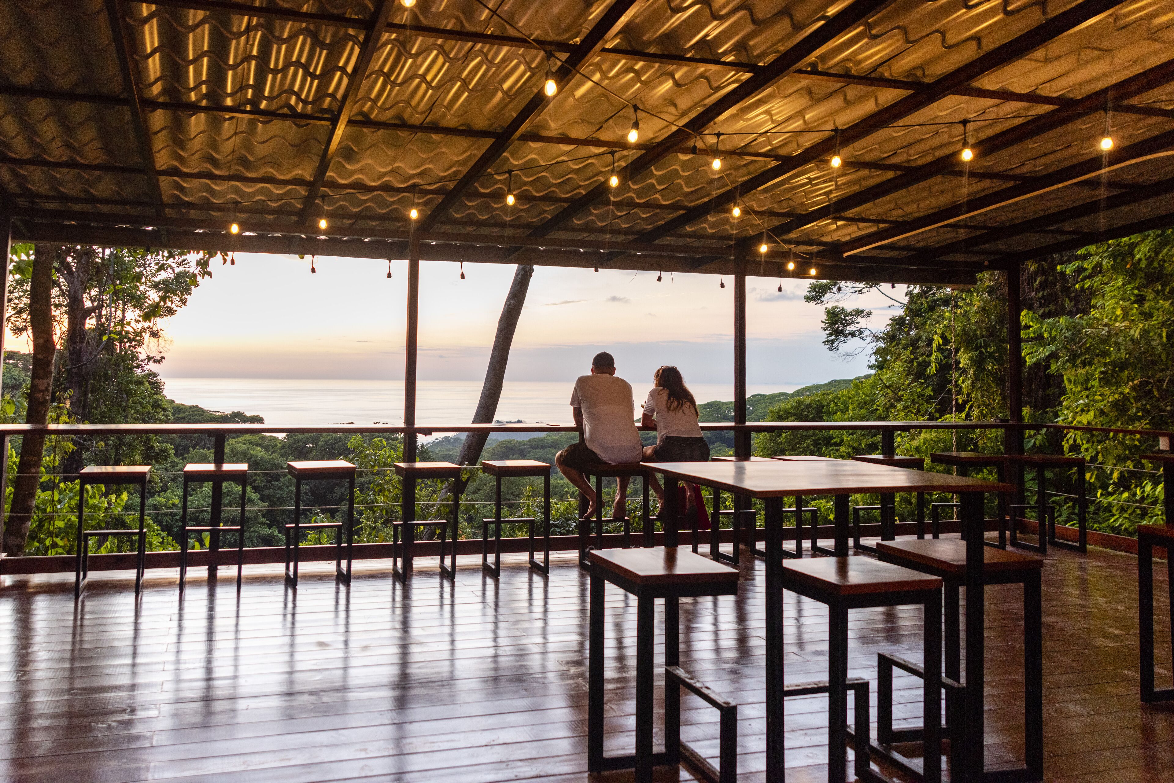 An outdoor seating area with wooden tables and benches, overlooking a scenic view of the ocean and hills.