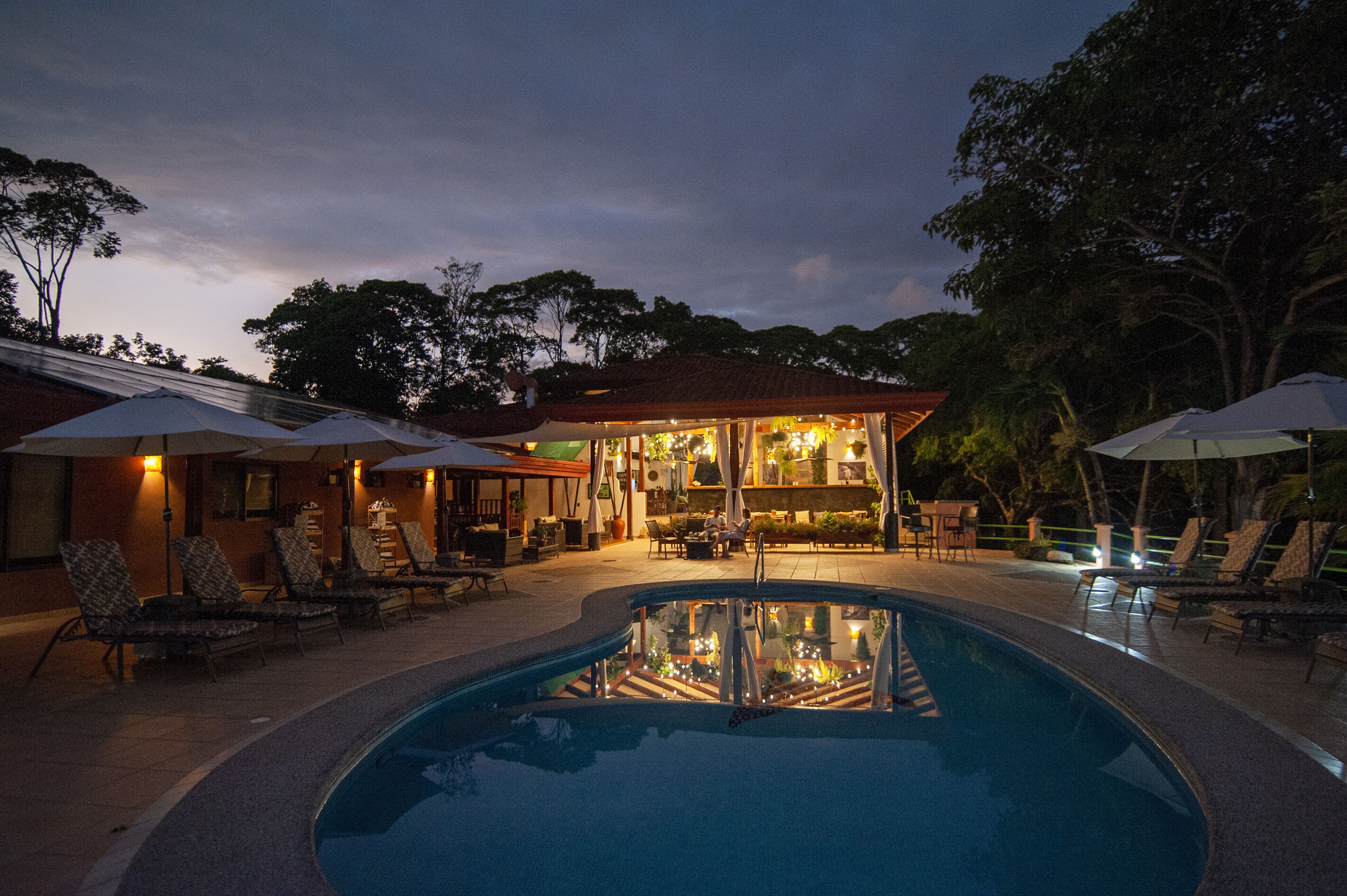 A pool area with lounge chairs and umbrellas, a bar, and a building with a red roof.
