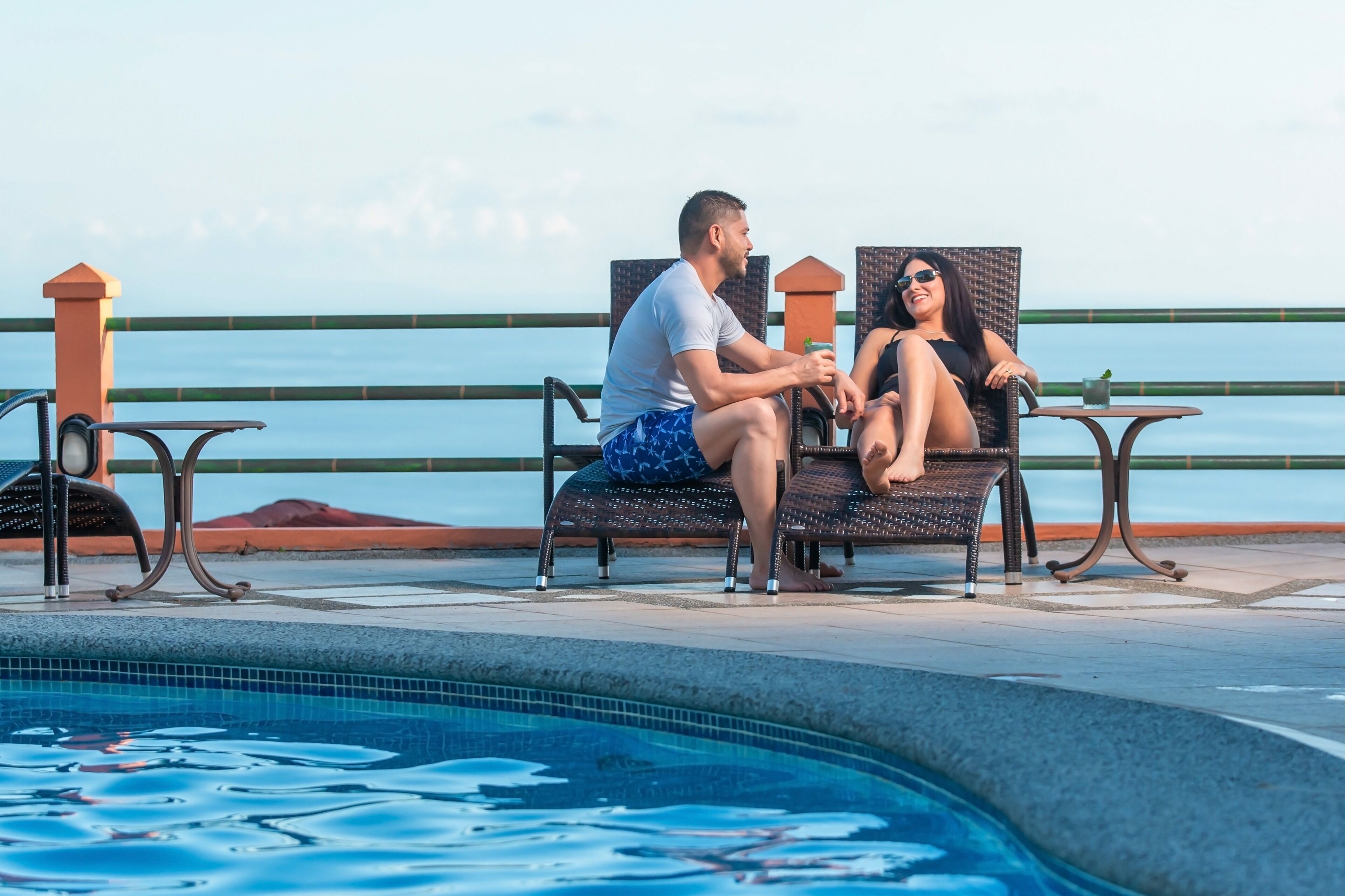 Un couple est assis sur des chaises longues au bord d’une piscine, avec vue sur la mer.