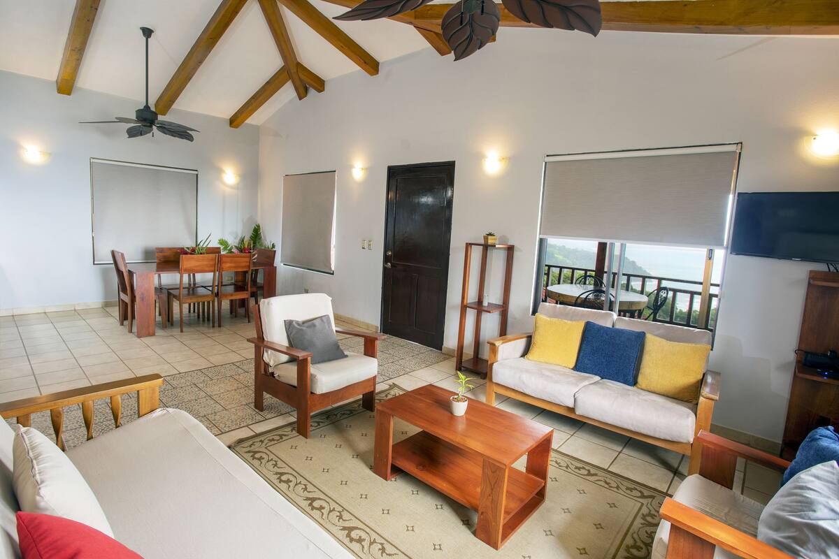 A spacious living room with a wooden ceiling, a dining area with a table and chairs, a sofa with cushions, and a television on the wall.