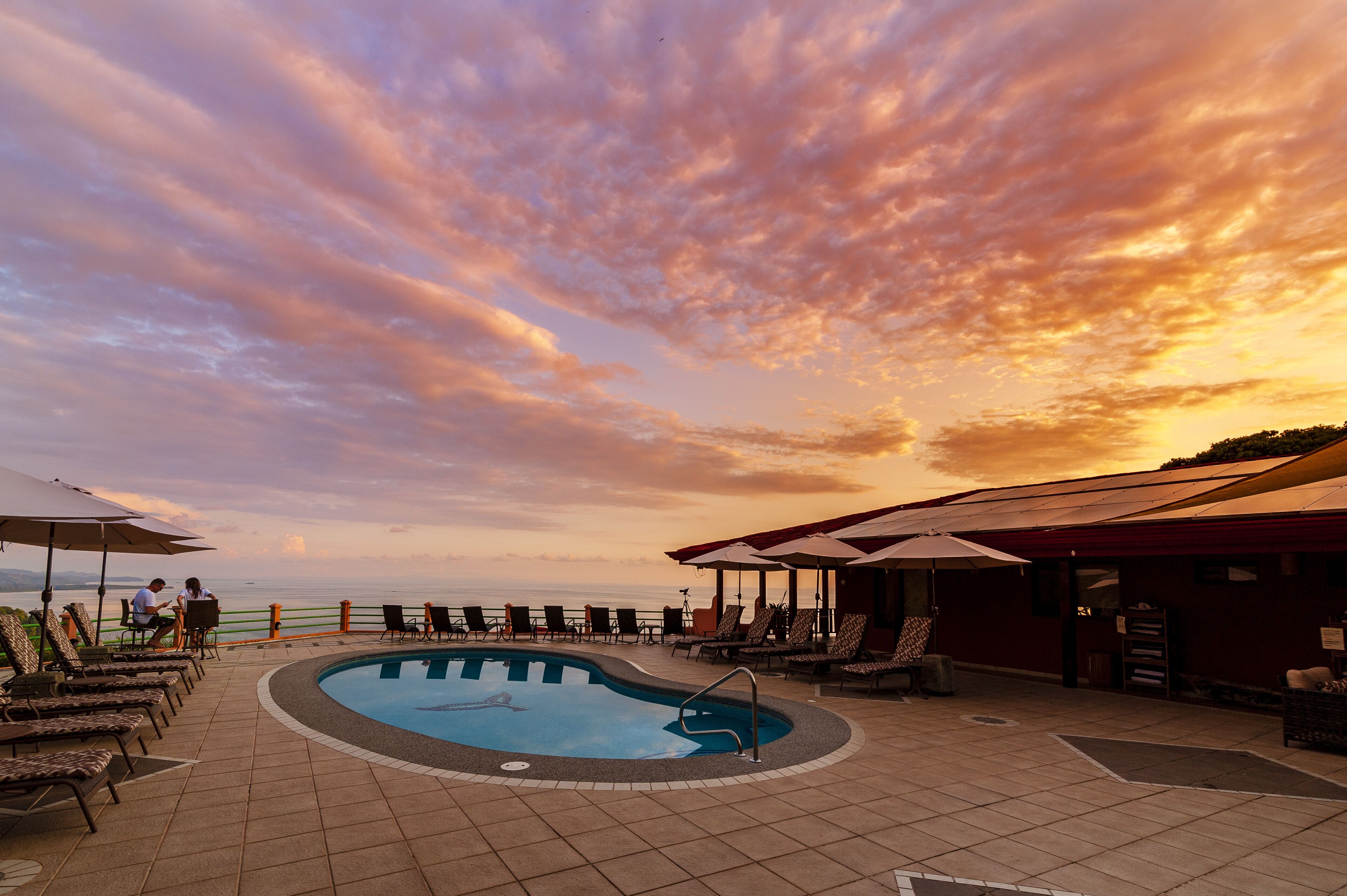 A poolside area with lounge chairs and umbrellas, overlooking a body of water during sunset.