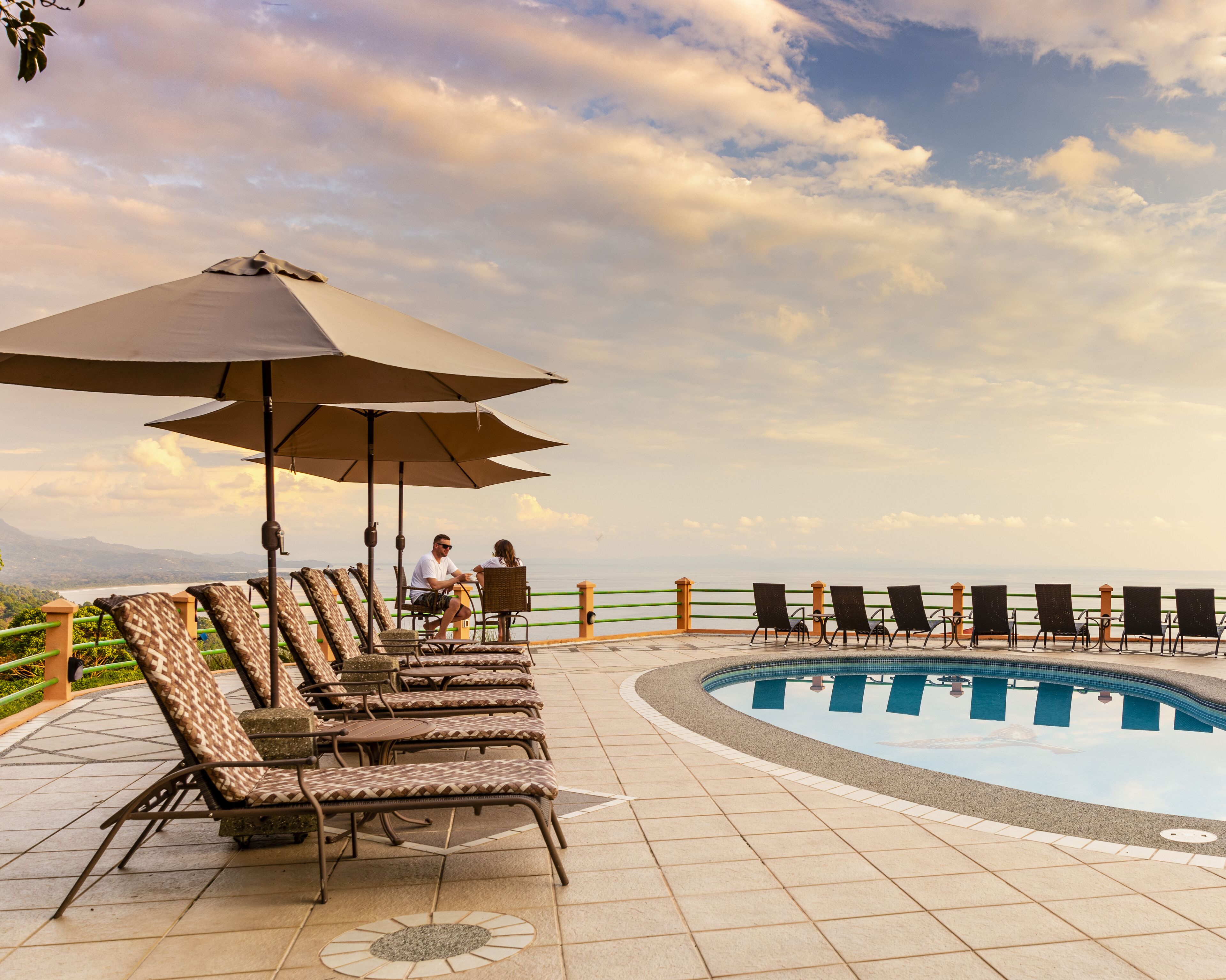 A poolside area with lounge chairs and umbrellas, a pool, and a view of the sea.
