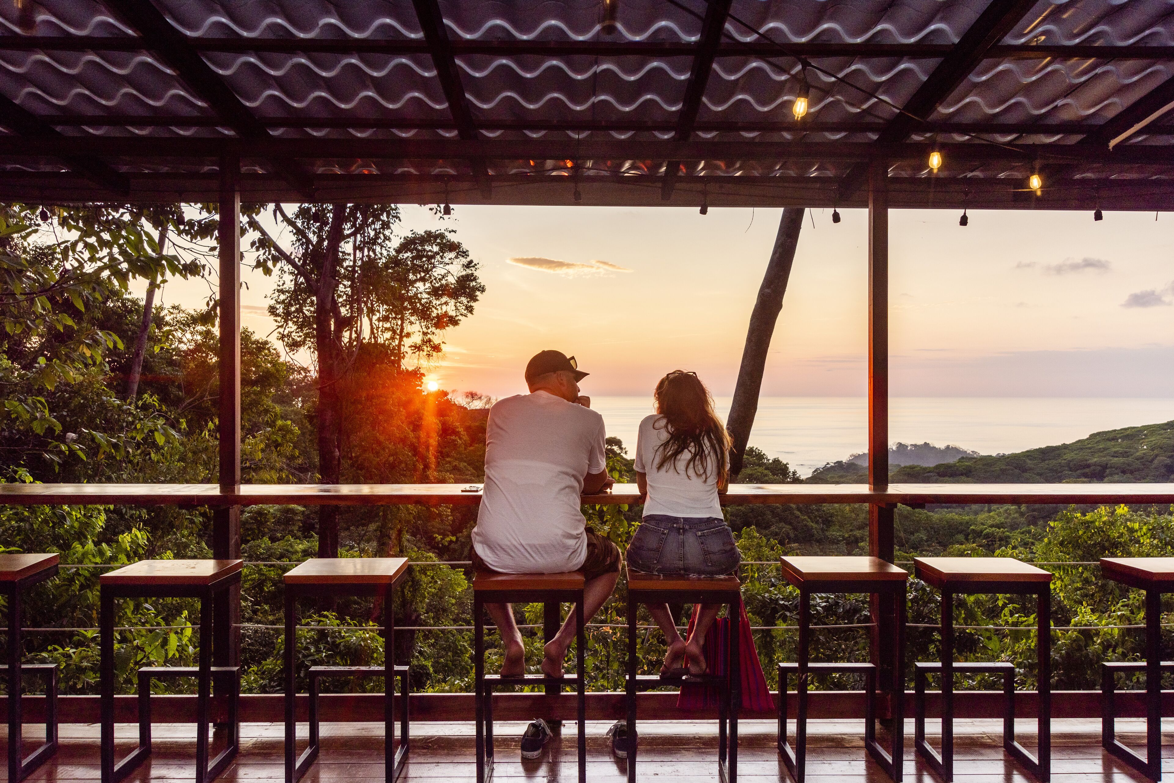 A couple sitting on a wooden bench overlooking a scenic view of a sunset and hills.