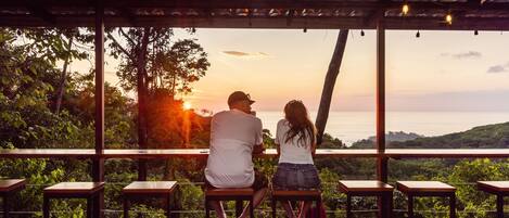 Una pareja sentada en un banco de madera contemplando una vista panorámica de una puesta de sol y colinas.