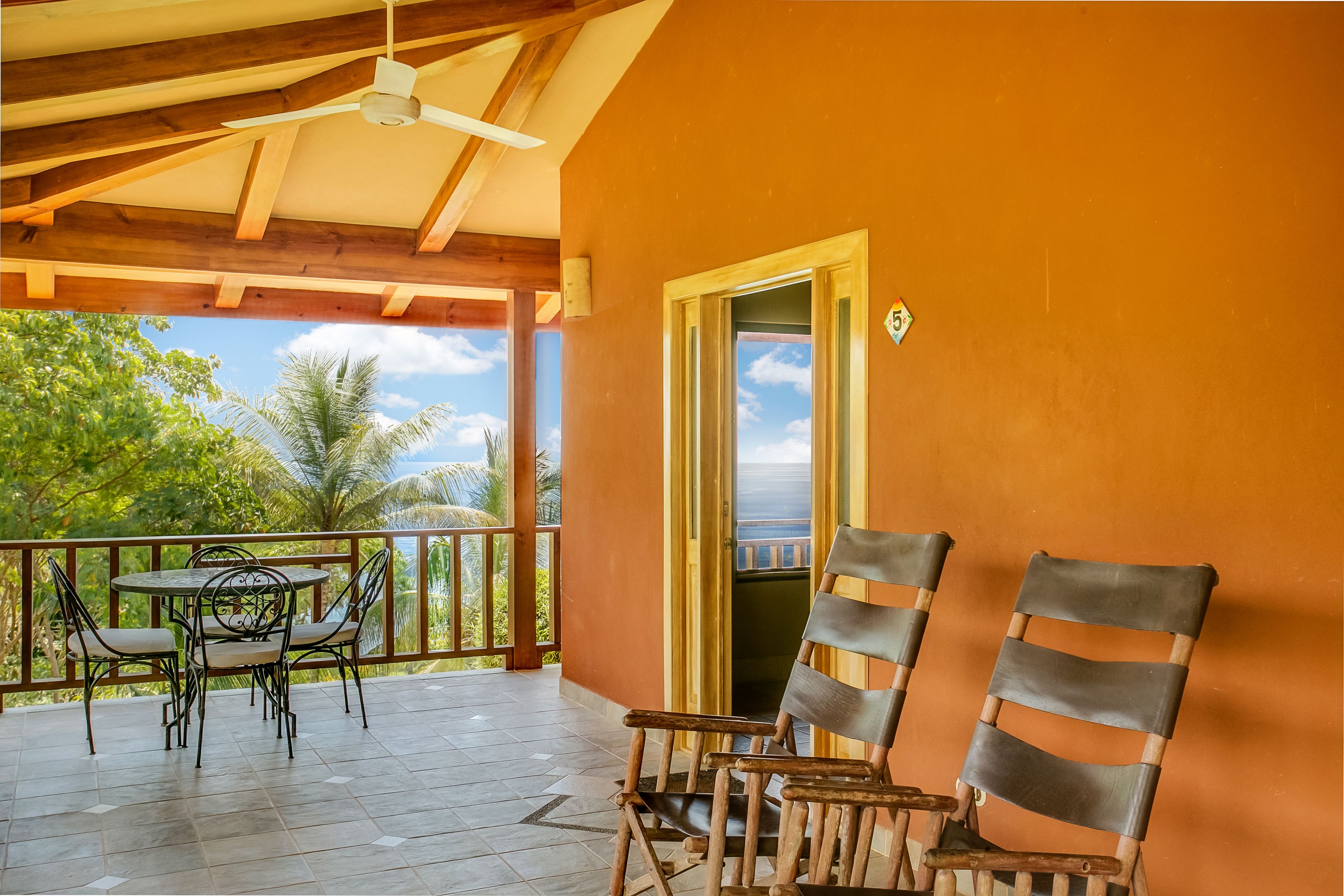 A covered outdoor area with a wooden ceiling, a table with chairs, and a view of palm trees and the ocean.