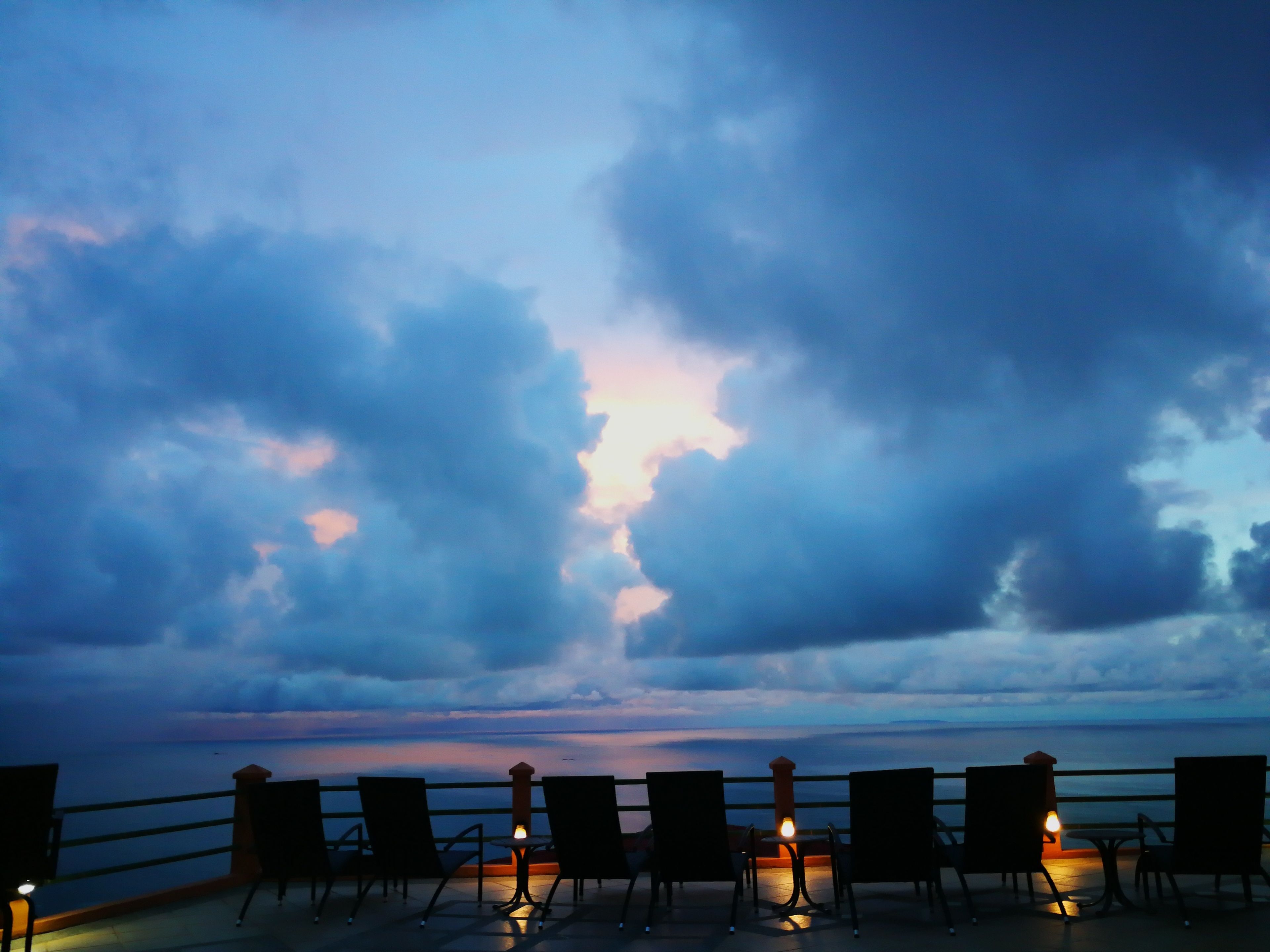 A terrace with chairs and tables, overlooking a body of water with a cloudy sky.