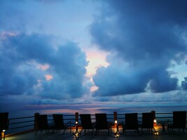 A terrace with chairs and tables, overlooking a body of water with a cloudy sky.
