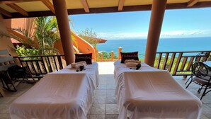 Two massage tables with white linens and pillows, a wooden chair, and a view of the ocean.
