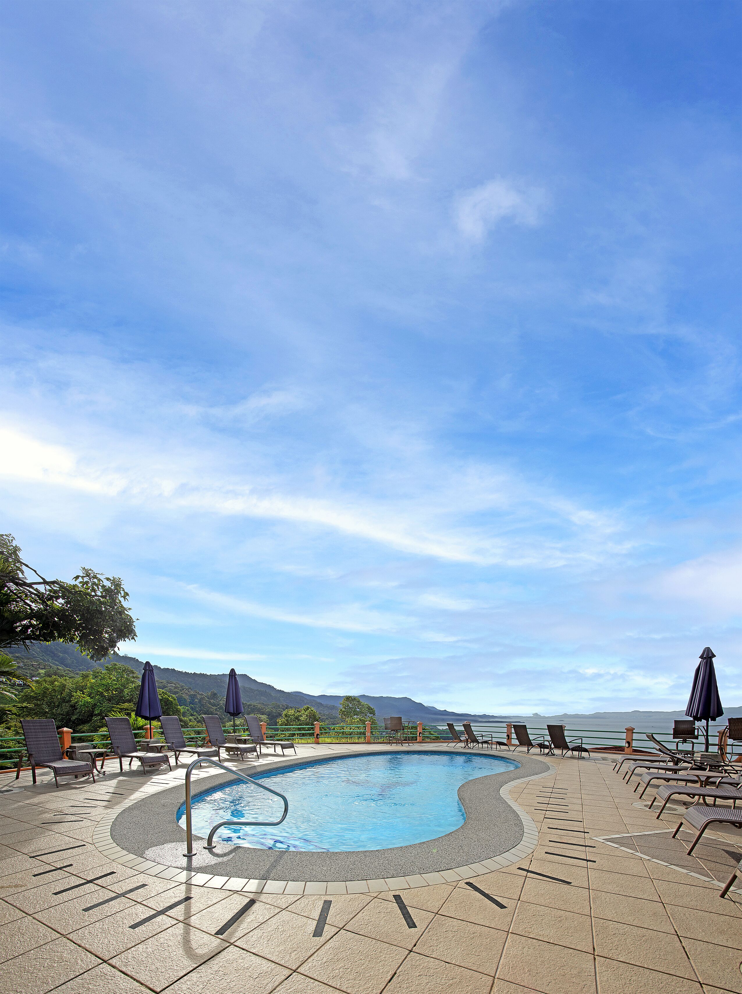 A pool area with lounge chairs and umbrellas, surrounded by a tiled deck and a view of the mountains.