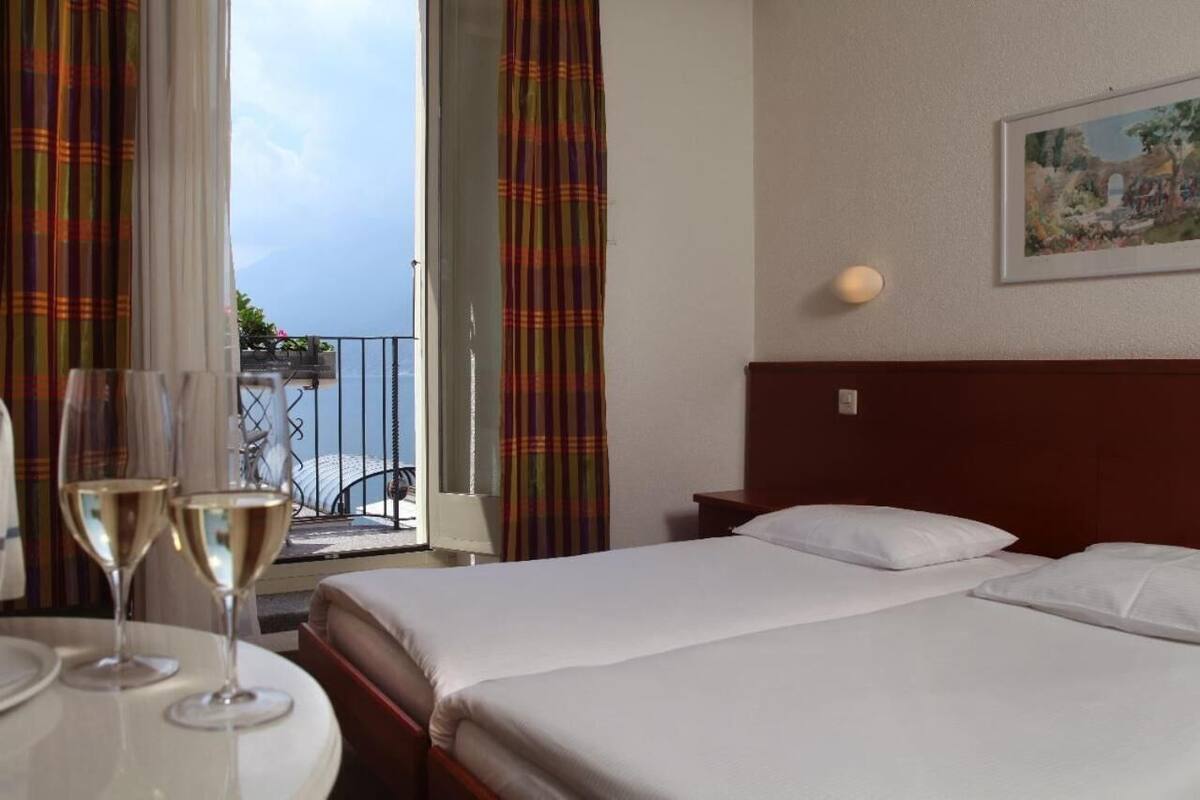 Une chambre d’hôtel avec un lit, une table sur laquelle reposent deux verres de vin, un tableau accroché au mur et une vue sur la mer par la fenêtre.