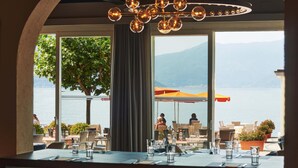A restaurant with outdoor seating overlooking a lake, featuring tables with chairs and a large window with a view of the water and mountains. - Piazza Ascona Hotel & Restaurants (Ascona)