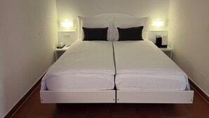 A double bed with white bedding and dark pillows, flanked by nightstands with lamps and a coffee maker. - Piazza Ascona Hotel & Restaurants (Ascona)