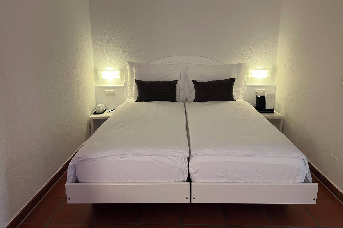 Un lit double avec une literie blanche et des oreillers foncés, de part et d’autre duquel se trouvent des tables de chevet avec des lampes et une machine à café.