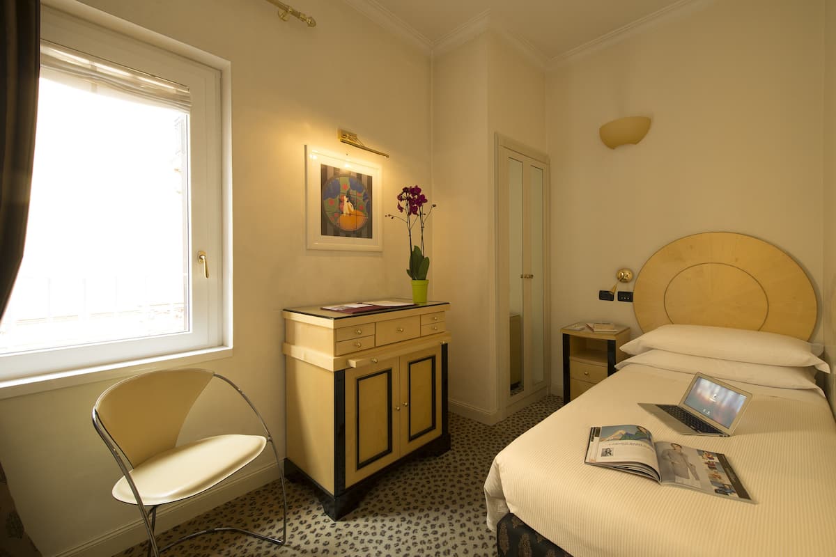 Habitación de hotel con cama, escritorio y silla, una mesita de noche con lámpara, un cuadro en la pared y ventana con cortinas.