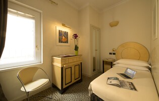 A hotel room with a bed, a desk with a chair, a small table with a lamp, a painting on the wall, and a window with curtains.
