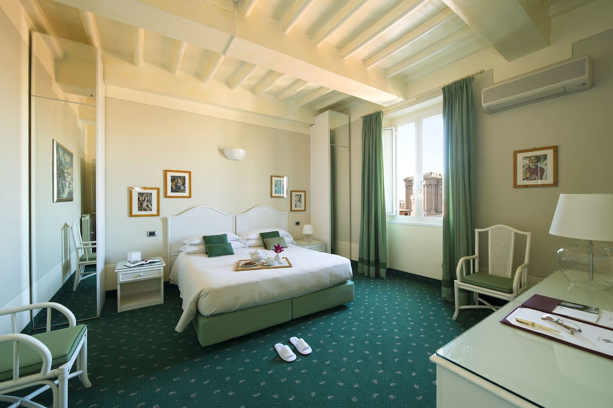 Una habitación de hotel con cama, escritorio, silla, ventana con cortinas e imágenes enmarcadas en las paredes.