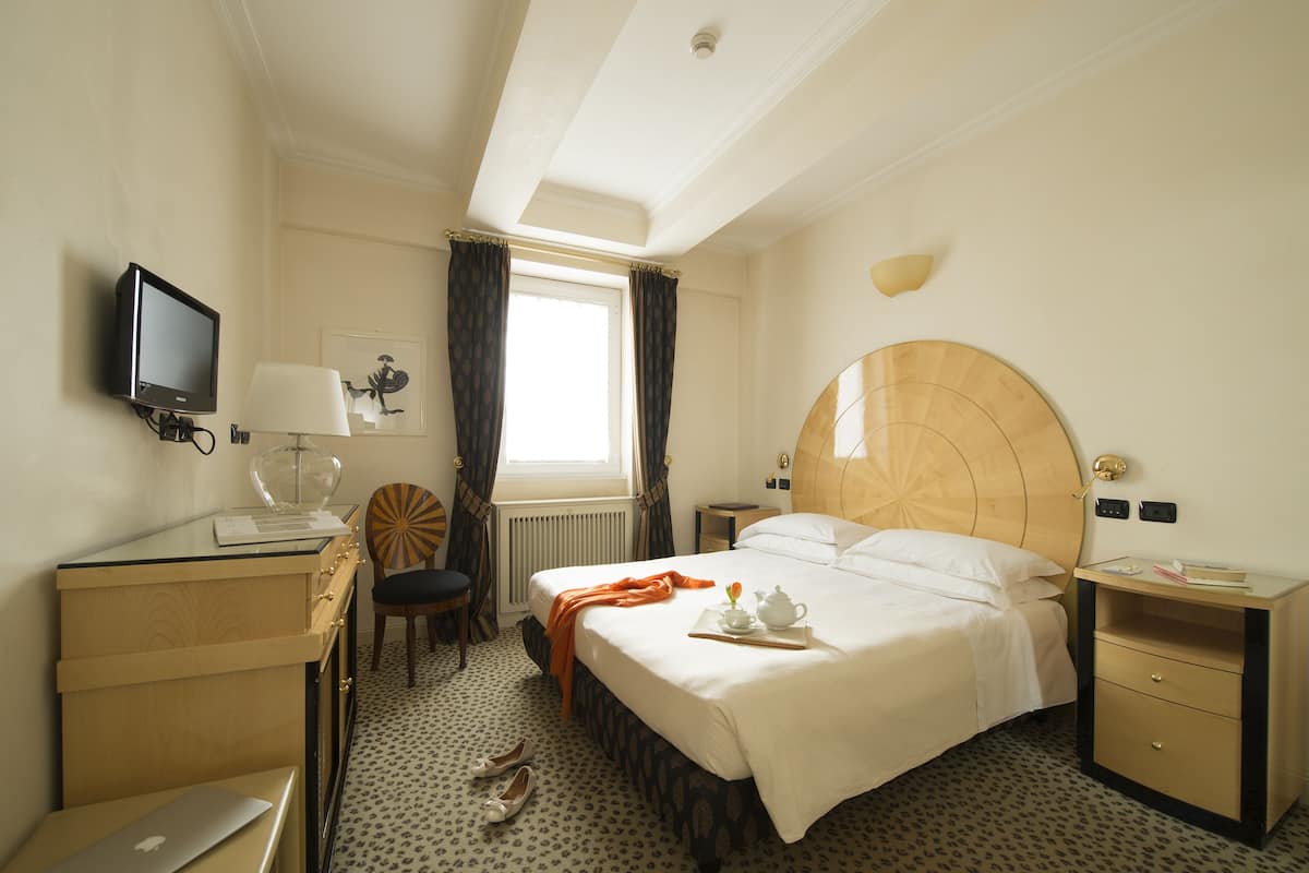 Habitación de hotel con cama, escritorio con computadora portátil, silla, televisor y ventana con cortinas.