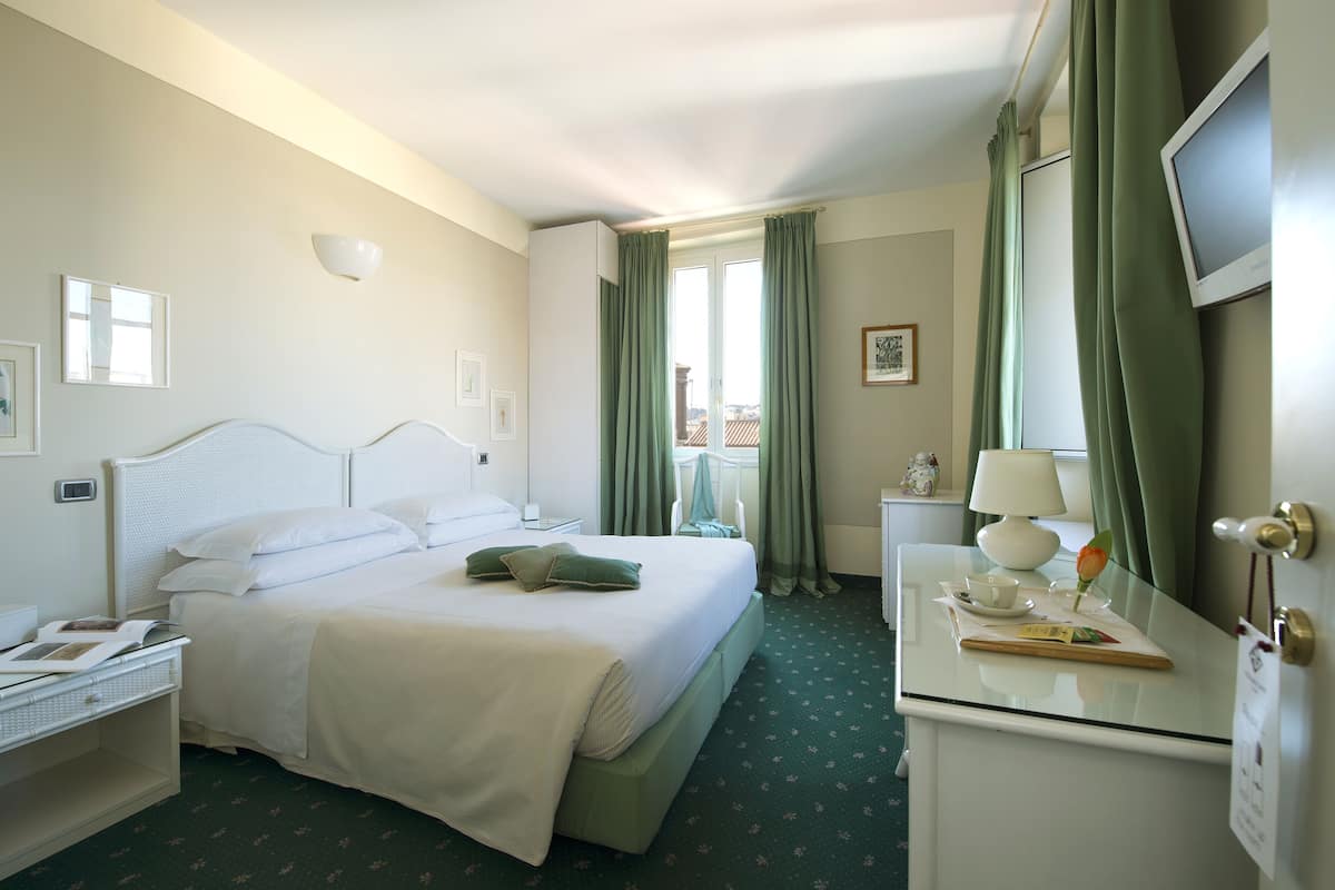 Habitación de hotel con cama, mesita de noche, lámpara y vistas al exterior a través de una ventana.