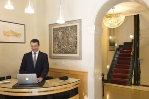A man at a reception desk with a laptop, framed artwork on the wall, and a staircase with red carpet.