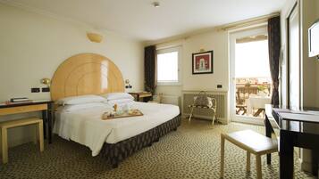 A hotel room with a large bed, a bedside table, a desk, a chair, a radiator, and a window with a view of the outside.