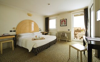 A hotel room with a large bed, a bedside table, a desk, a chair, a radiator, and a window with a view of the outside.