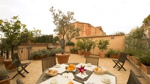 Un patio exterior con una mesa preparada para el desayuno, rodeado de plantas en macetas y vistas a un edificio.