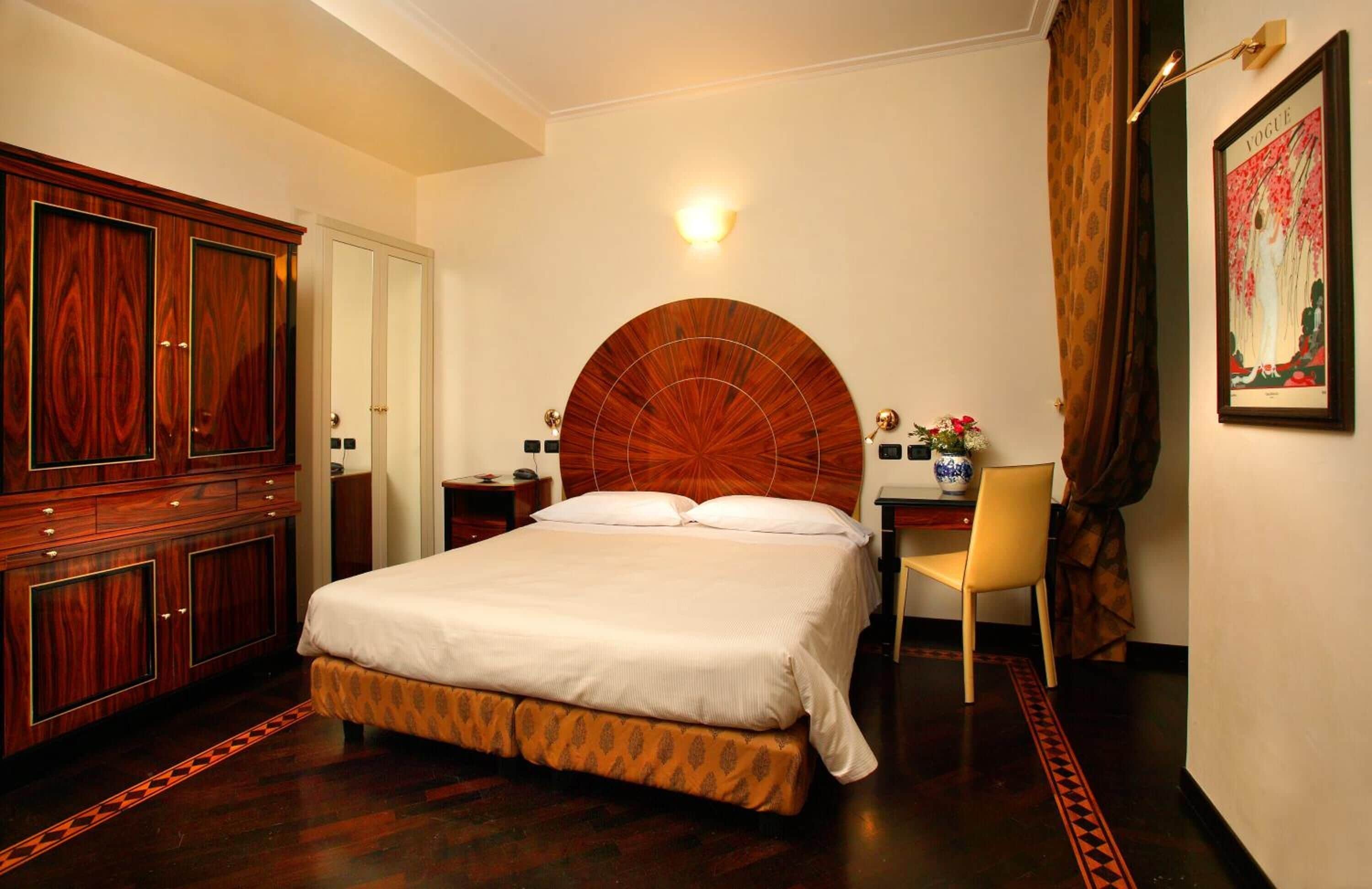 A bedroom with a large bed, wooden wardrobe, a desk with a chair, and a wall-mounted light fixture.