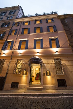 Een hotelgevel van steen, met meerdere ramen voorzien van luiken, en een goed verlichte entree.