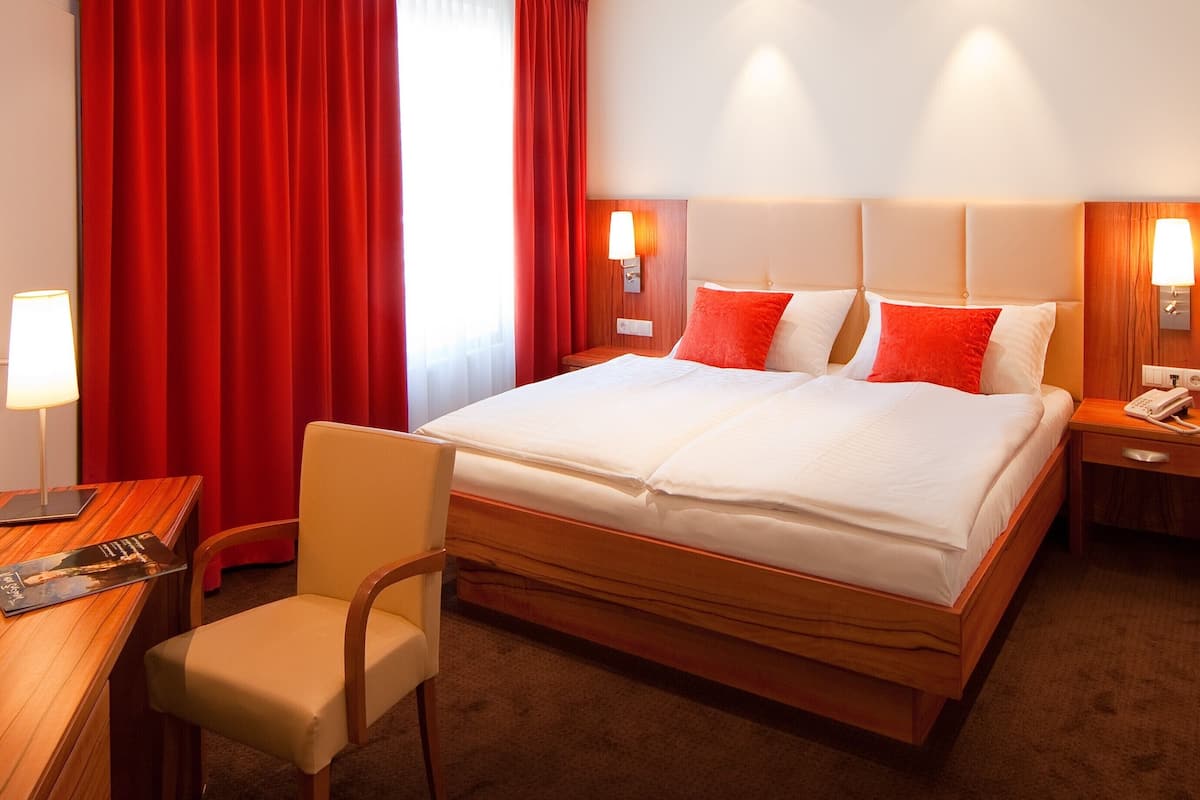 Ein Hotelzimmer mit einem großen Bett, zwei Nachttischlampen, einem Schreibtisch mit Stuhl und roten Vorhängen.