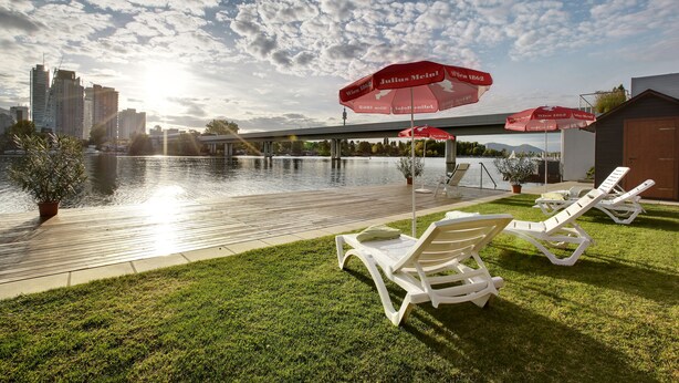 A poolside area with lounge chairs and umbrellas, a city skyline in the background, and a clear sky. - Strandhotel Alte Donau (Vienna)