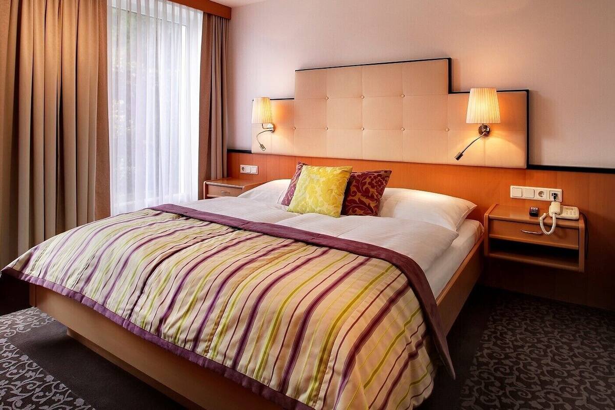 Ein Hotelzimmer mit einem großen Bett, gestreifter Bettwäsche, zwei Nachttischlampen, einem Nachttisch mit Telefon und einem Fenster mit Vorhängen.