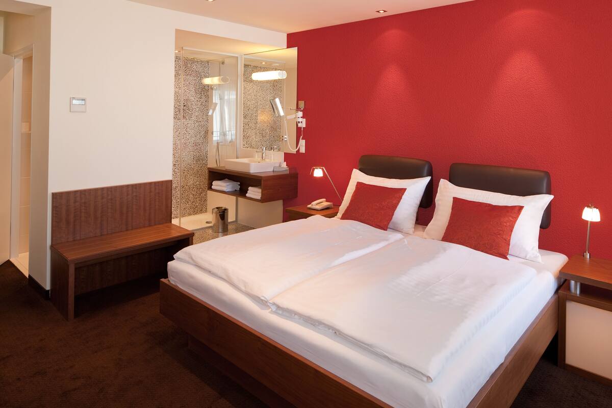 Ein Hotelzimmer mit einem großen Bett, einer Holzbank, einem Schminktisch mit Spiegel und einer roten Akzentwand.