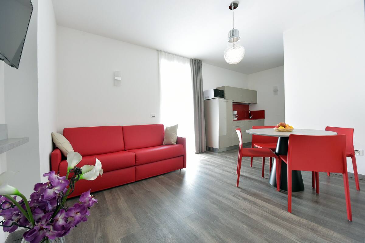 Ein modernes Wohnzimmer mit einem roten Sofa, einem Esstisch mit roten Stühlen und einer Küche im Hintergrund.