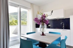 Una cocina moderna con armarios blancos y un salpicadero oscuro, un comedor con mesa y sillas, y vistas a la vegetación a través de grandes ventanales.