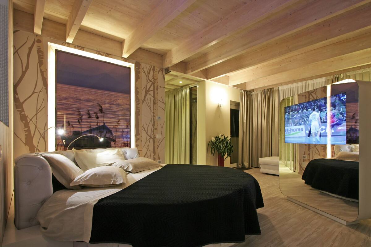 Ein modernes Hotelzimmer mit einem großen Bett, einem Flachbildfernseher, einem großen Fenster mit Sonnenuntergangsblick und einer Holzbalkendecke.
