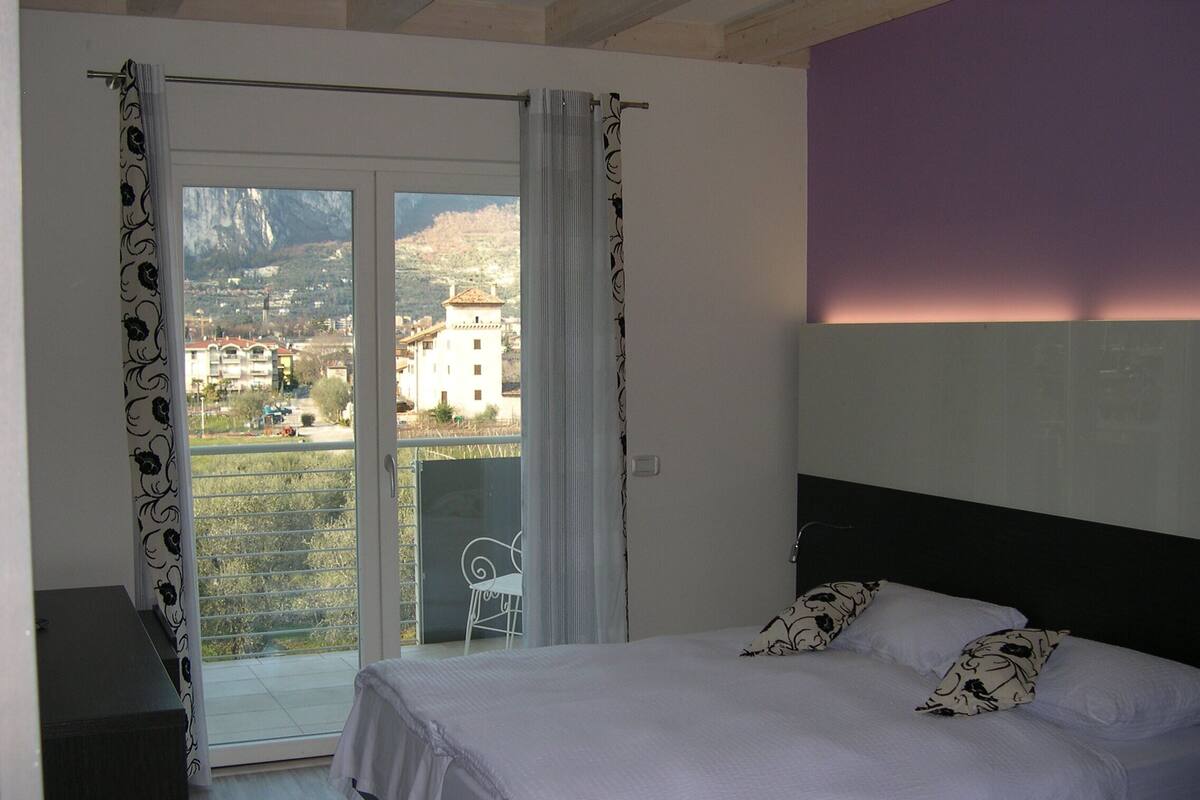 Ein Schlafzimmer mit Bett, Blick auf eine gebirgige Landschaft durch eine Schiebetür und einem Balkon mit einem Stuhl.