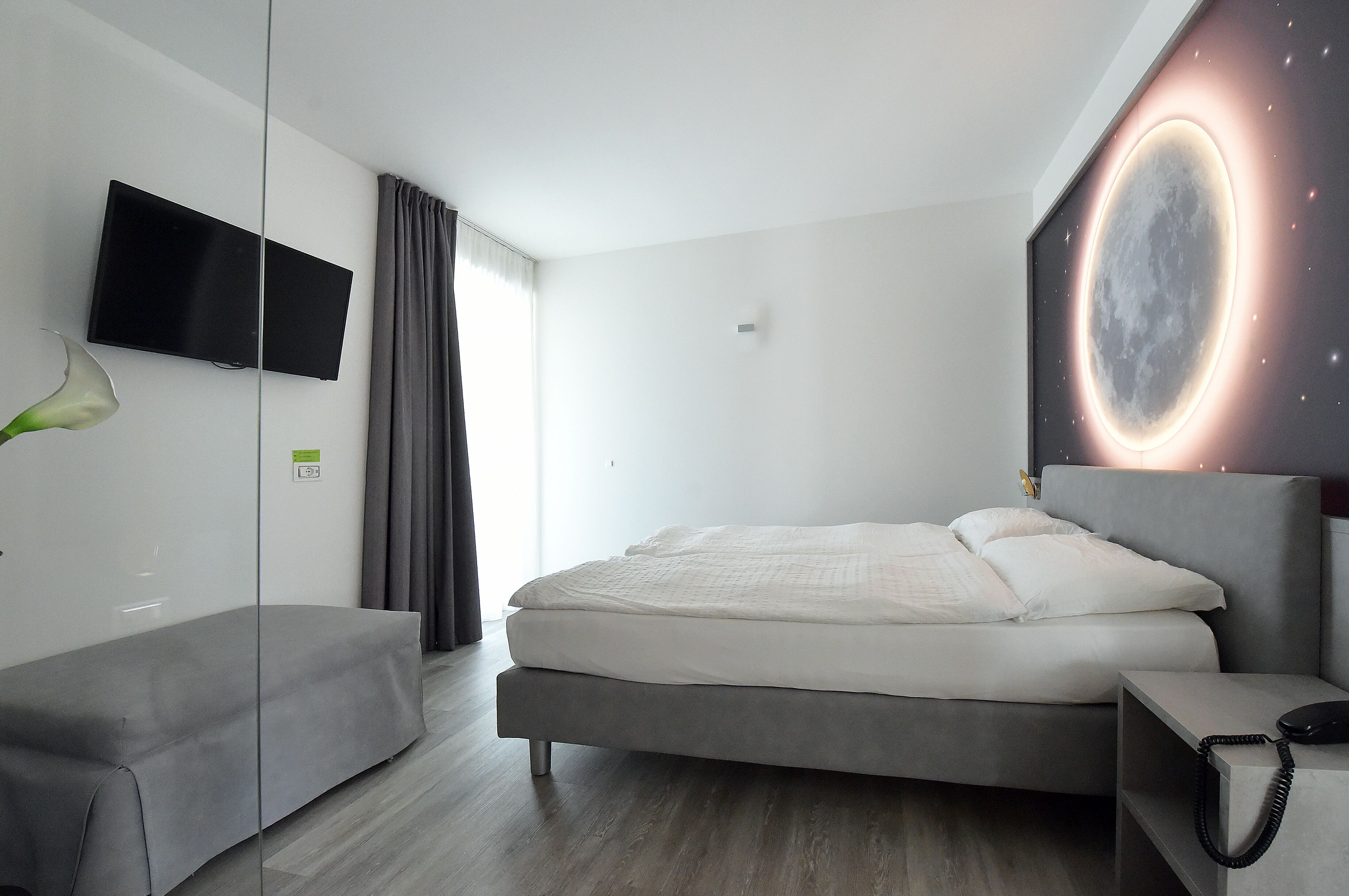 A modern hotel room with a bed, bedside table with a phone, a flat-screen TV mounted on the wall, and a large window with curtains.