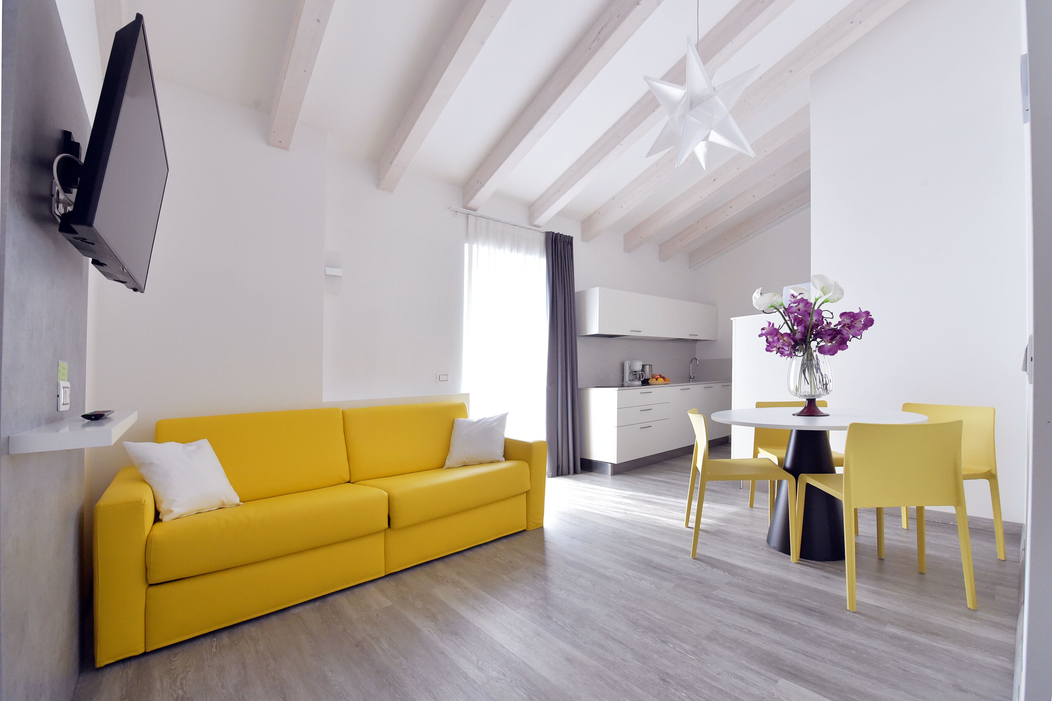 Ein modernes Wohnzimmer mit einem gelben Sofa, einem Essbereich mit gelben Stühlen, einer Küche im Hintergrund und einem Wand-Fernseher.