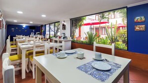 A restaurant with white tables and yellow chairs, a large window with a view of the outside, and a blue wall with two paintings. - La Cartuja (Quito)