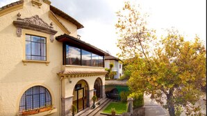 A two-story building with a large bay window, arched doorways, and a small garden area with a tree and potted plants. - La Cartuja (Quito)