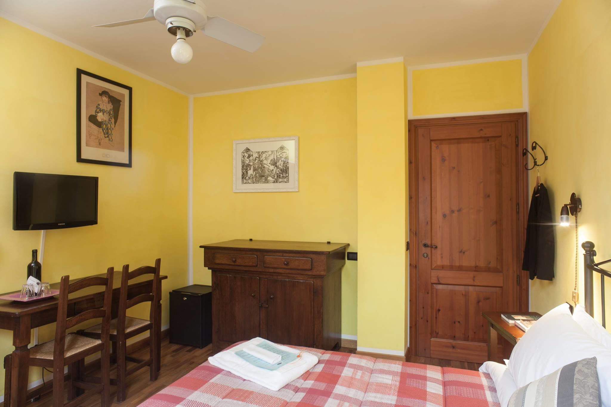 A room with yellow walls, a wooden wardrobe, a bed with a checkered bedspread, a desk with a chair, a television, and a ceiling fan.