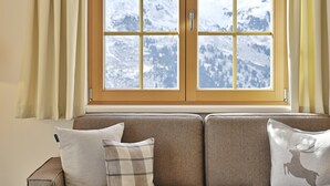 A window with a view of snow-covered mountains, a sofa with two cushions, and a wooden side table. - AlpinLodges Kühtai (Silz)