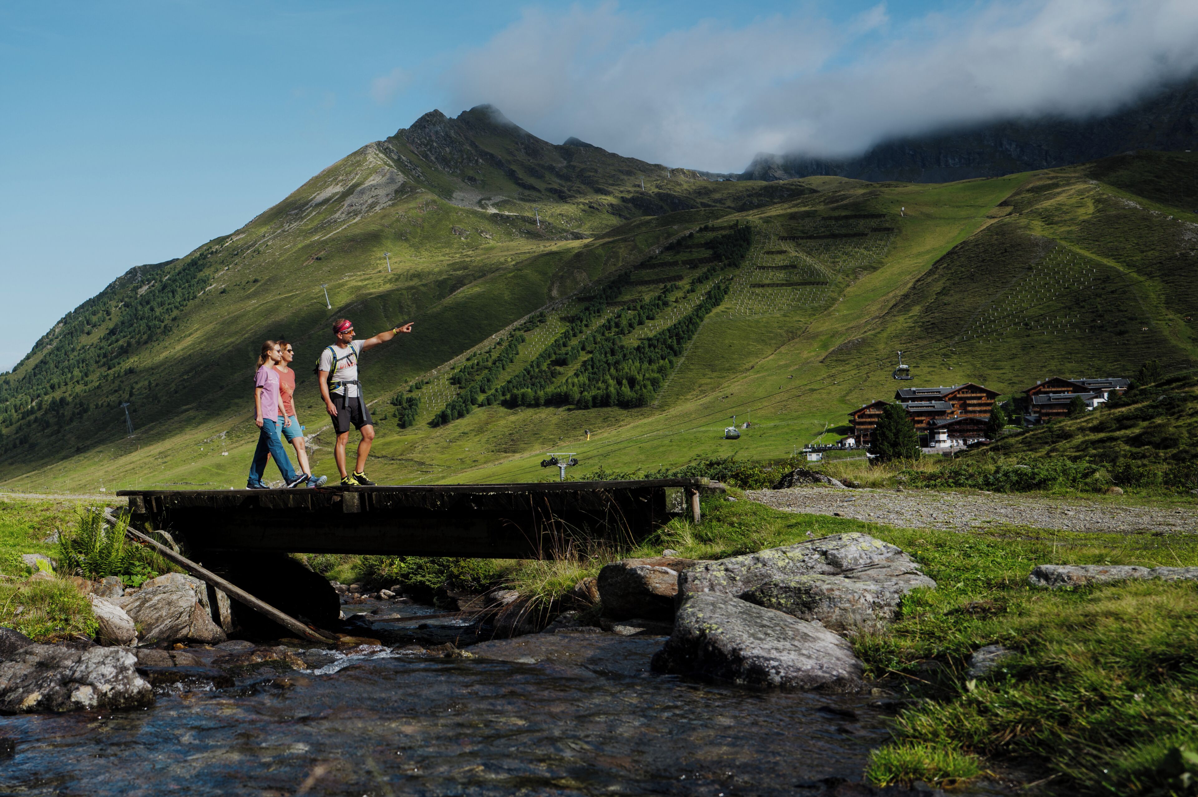 A bridge over a stream with a mountainous backdrop and a building with a sloped roof.